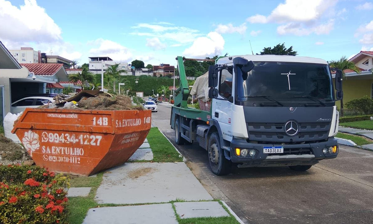 Caçamba laranja da S.A. Entulho cheia de entulho na calçada de um condomínio, com a marca e os dois telefones pintados em branco na lateral, e o caminhão chegando para a retirada.